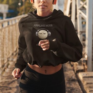 May include: Black cropped hoodie with the text "AIRPLANE MODE" and a graphic of a grumpy, fluffy ball wearing headphones. The model is wearing black leggings and pink headphones.