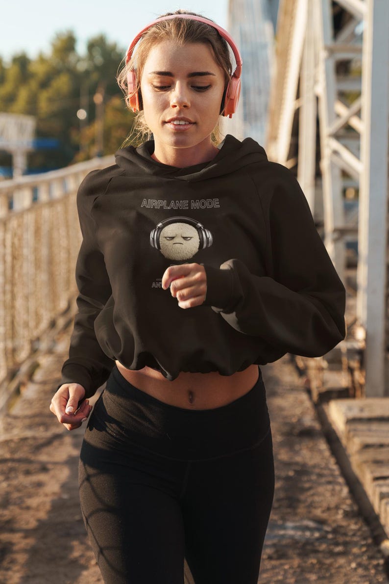 May include: Black cropped hoodie with the text "AIRPLANE MODE" and a graphic of a grumpy, fluffy ball wearing headphones. The model is wearing black leggings and pink headphones.