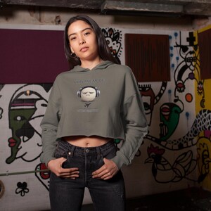 May include: A cropped olive green hoodie with a graphic of a face wearing headphones and the text "EVERYTHING IS ABOUT". The model is wearing black jeans. The background is a graffiti-covered wall.