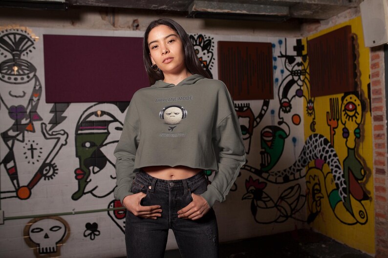 May include: A cropped olive green hoodie with a graphic of a face wearing headphones and the text "EVERYTHING IS ABOUT". The model is wearing black jeans. The background is a graffiti-covered wall.