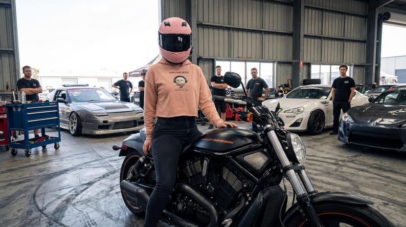 May include: A person wearing a pink helmet and a cropped peach hoodie with a graphic, sitting on a black Harley-Davidson motorcycle. Several cars are in the background, including a silver sports car and a white sports car. The setting appears to be a garage or workshop.