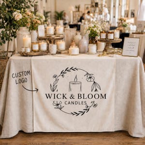 Op de afbeelding: Een beige linnen tafelloper met een zwart gedrukt logo met de tekst WICK & BLOOM CANDLES, gecentreerd in een bloemenkransafbeelding. De loper ligt op een tafel vol met diverse glazen kaarsen, vazen en bloemstukken.