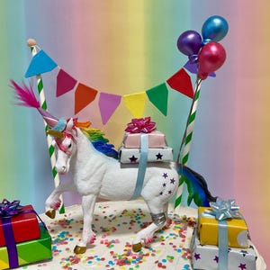 May include: A whimsical cake topper featuring a white unicorn with a rainbow mane and tail, carrying gifts. The unicorn is adorned with a party hat and surrounded by colorful balloons and a banner. The gifts are wrapped in red, green, pink, and yellow boxes.
