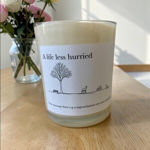 Puede incluir: Vela perfumada en un recipiente de vidrio transparente. La etiqueta dice "A life less hurried" con un dibujo de un &aacute;rbol, un banco y ciervos. La vela est&aacute; sobre una superficie de madera. Un jarr&oacute;n con flores est&aacute; en el fondo.