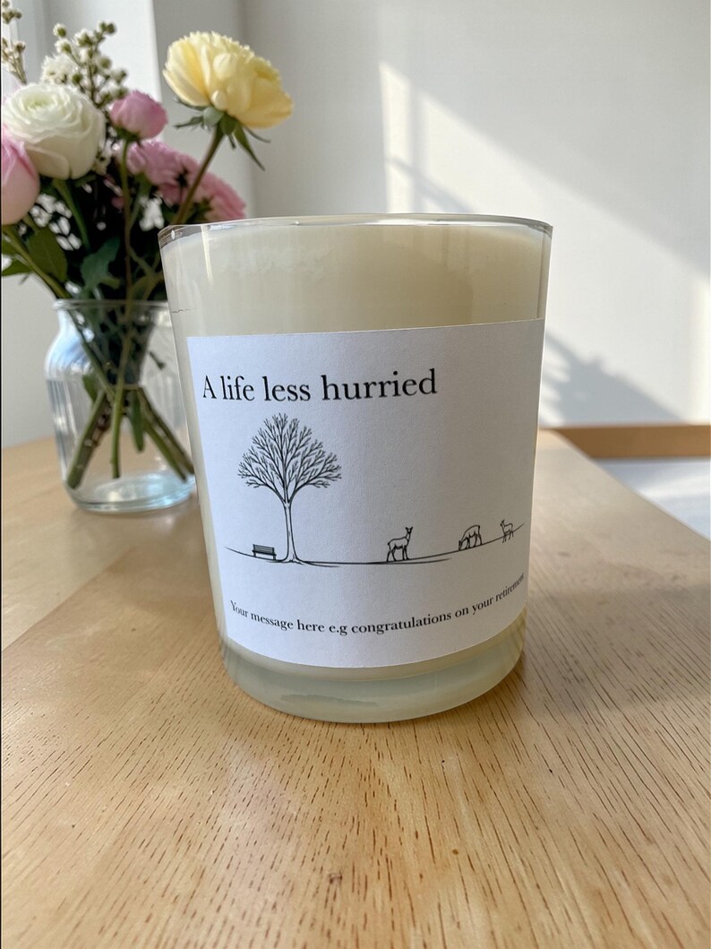 Puede incluir: Vela perfumada en un recipiente de vidrio transparente. La etiqueta dice "A life less hurried" con un dibujo de un &aacute;rbol, un banco y ciervos. La vela est&aacute; sobre una superficie de madera. Un jarr&oacute;n con flores est&aacute; en el fondo.