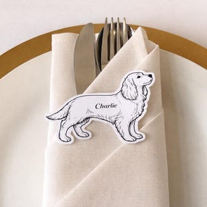May include: A black and white dog-shaped place card with the name "Charlie" printed on it, resting on a folded beige napkin. Silverware, including a fork and knife, is tucked into the napkin. The place setting is on a white plate with a gold rim.