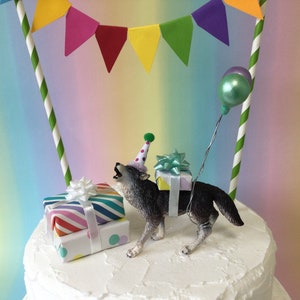 May include: A gray wolf cake topper wearing a polka dot party hat and carrying a gift box and a balloon. The wolf is standing on a white cake with a colorful bunting behind it.