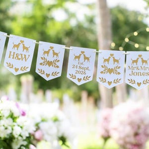 May include: A white banner with gold accents, strung together, featuring dog silhouettes, hearts, and text. Some banners display initials, a wedding date, and the names "Mr & Mrs Jones". The banner is set against a blurred green background.
