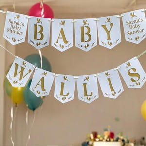 May include: White banner with gold lettering spelling "BABY WALLIS" and "Sarah's Baby Shower". The banner is decorated with bird and footprint motifs. Balloons in pink, yellow, and teal are visible in the background.