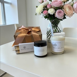 Puede incluir: Un juego de regalo con una caja marr&oacute;n atada con una cinta dorada, una caja de cerillas, un tarro negro con la etiqueta "SPA RECOVERY" y una vela con el texto "A life less hurried". Un jarr&oacute;n de flores rosas y blancas est&aacute; en el fondo.