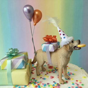 May include: A yellow Labrador Retriever dog cake topper wearing a white and red polka dot party hat with a feather, holding a yellow cookie. The dog is carrying a light blue and purple striped gift box with an orange bow. There are two balloons, one purple and one orange, and a stack of two gift boxes, one light blue and purple striped and one light green, on the cake.