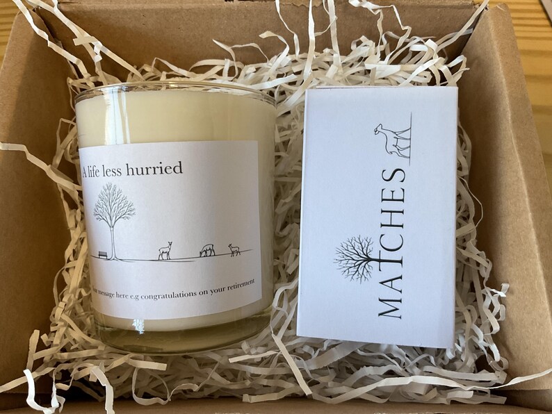 Puede incluir: Un juego de regalo en una caja de cart&oacute;n, que contiene una vela de vidrio con el texto "A life less hurried" y una peque&ntilde;a caja blanca de cerillas. La vela tiene una etiqueta con una ilustraci&oacute;n de un &aacute;rbol y ciervos. La caja de cerillas dice "MATCHES".