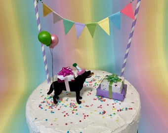 May include: A birthday cake with a black dog figurine wearing a party hat and carrying gifts. The cake is decorated with colorful sprinkles, balloons, and a banner. A second gift box is also on the cake.