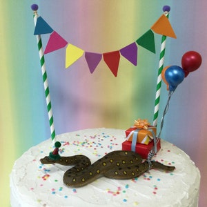 Könnte beinhalten: Ein weißer Kuchen mit einer braunen Schlange, die einen Partyhut trägt, einer bunten Flaggenkette und Geschenken auf der Oberseite. Der Kuchen ist mit bunten Streuseln verziert.