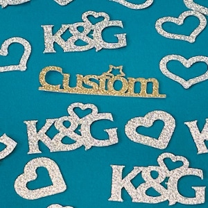 May include: Glittery silver K&G and heart-shaped cutouts scattered on a teal background. A gold glitter cutout with the word "Custom" is also present. These decorative elements are ideal for crafting and embellishments.