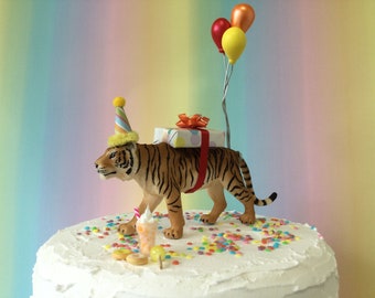 May include: A white cake with a tiger figurine wearing a party hat and carrying a wrapped gift. The tiger is standing on a bed of colorful sprinkles. There are three balloons tied to the gift, one red, one orange, and one yellow.