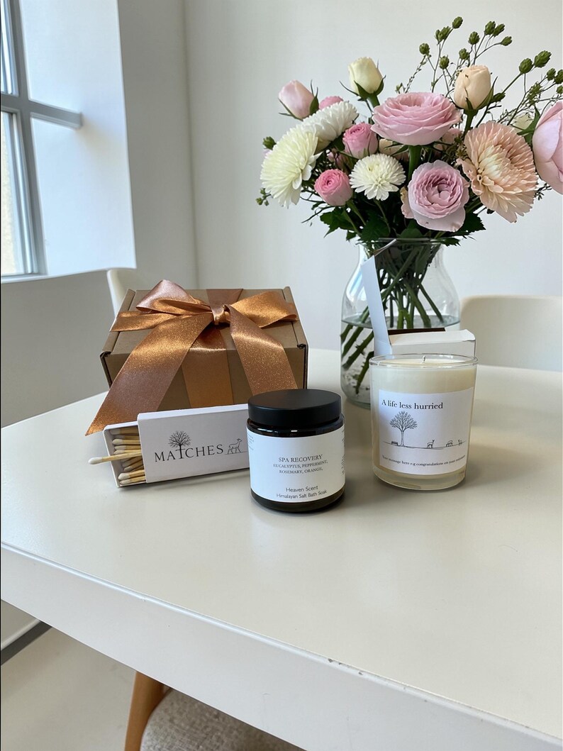 Puede incluir: Un bodeg&oacute;n con un ramo de flores rosas y blancas en un jarr&oacute;n de cristal, una caja de regalo marr&oacute;n con una cinta dorada, un tarro de sal de recuperaci&oacute;n spa, una caja de cerillas y una vela con la frase "A life less hurried".