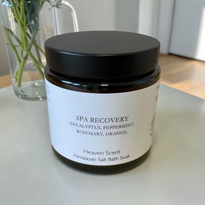 Puede incluir: Un tarro de vidrio oscuro con tapa negra y etiqueta blanca. La etiqueta dice "SPA RECOVERY" con los ingredientes: eucalipto, menta, romero y naranja. Es una sal de ba&ntilde;o del Himalaya de Heaven Scent.