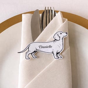 May include: A white dachshund-shaped place card with the name "Danielle" in script, placed on a folded beige napkin with silverware. The napkin is on a white plate with a gold rim, suggesting a wedding or event setting.