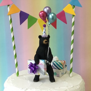 May include: Black bear cake topper wearing a party hat and holding balloons and a gift box. The bear is sitting on a white cake with green and white striped straws and colorful paper flags.