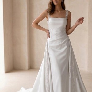 Pode incluir: Um vestido de noiva branco com decote quadrado e al&ccedil;as adornadas. O vestido apresenta um corpete ajustado e uma saia fluida com cauda. Detalhes delicados de contas adornam as laterais da saia, adicionando um toque de brilho ao design elegante.