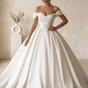May include: A white off-the-shoulder wedding gown with a fitted bodice and a full, flowing skirt. The dress features a sweetheart neckline and a defined waist. The fabric appears to be a smooth, satin-like material. The gown is elegant and classic.