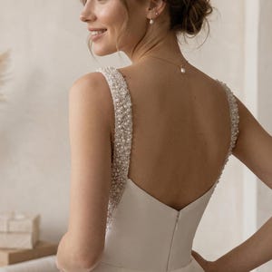 Pode incluir: Um vestido de noiva branco com decote em V e al&ccedil;as com contas. O vestido tem um corpete justo e uma saia fluida. As al&ccedil;as s&atilde;o adornadas com pequenas contas brilhantes. As costas do vestido apresentam um corte em V baixo e um fecho de correr.