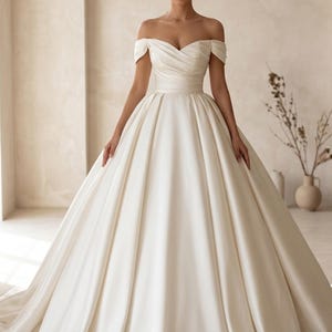 May include: An off-white, off-the-shoulder wedding gown with a full skirt. The bodice features a sweetheart neckline and a cinched waist. The dress is made of a smooth, satin-like fabric. The gown is floor-length and has a slight train.