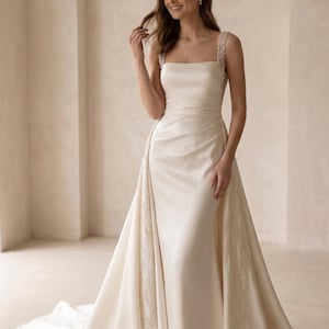Pode incluir: Vestido de noiva marfim com decote quadrado e cauda fluida. O vestido apresenta al&ccedil;as adornadas e um corpete justo que se abre em uma saia ampla. O design do vestido &eacute; cl&aacute;ssico e elegante, adequado para um casamento formal.