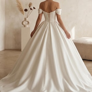 May include: A white off-the-shoulder wedding gown with a ball gown silhouette. The dress features a lace-up back and a long, flowing skirt. The model is turned away from the camera, showcasing the back of the dress.