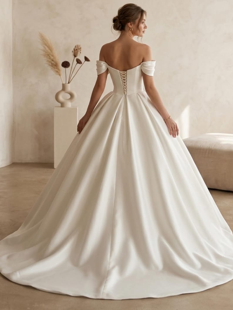 May include: A white off-the-shoulder wedding gown with a ball gown silhouette. The dress features a lace-up back and a long, flowing skirt. The model is turned away from the camera, showcasing the back of the dress.