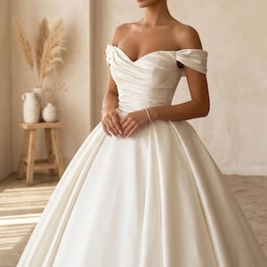 May include: A white satin off-the-shoulder wedding gown with a sweetheart neckline and a full skirt. The dress features a fitted bodice and a flowing skirt, creating an elegant silhouette. The gown is designed for a formal occasion.