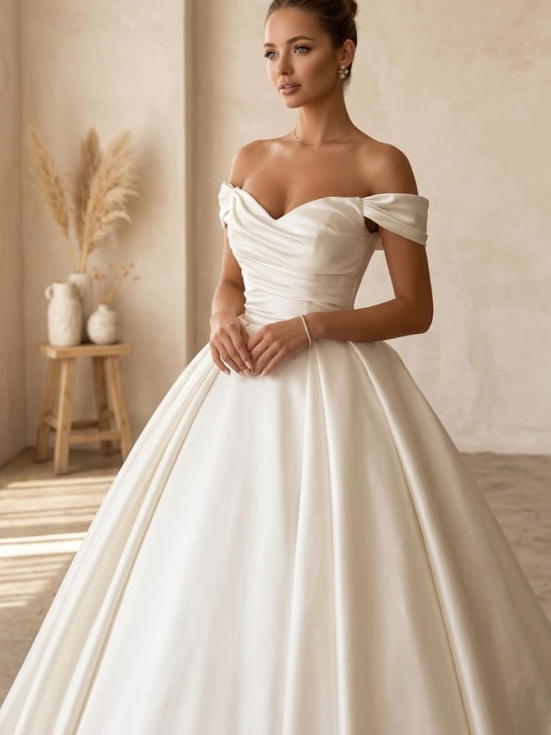 May include: A white satin off-the-shoulder wedding gown with a sweetheart neckline and a full skirt. The dress features a fitted bodice and a flowing skirt, creating an elegant silhouette. The gown is designed for a formal occasion.