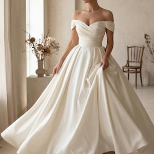 May include: An off-white, off-the-shoulder wedding gown with a full skirt. The dress features a sweetheart neckline and a fitted bodice that flows into a voluminous skirt. The bride is wearing white heels.