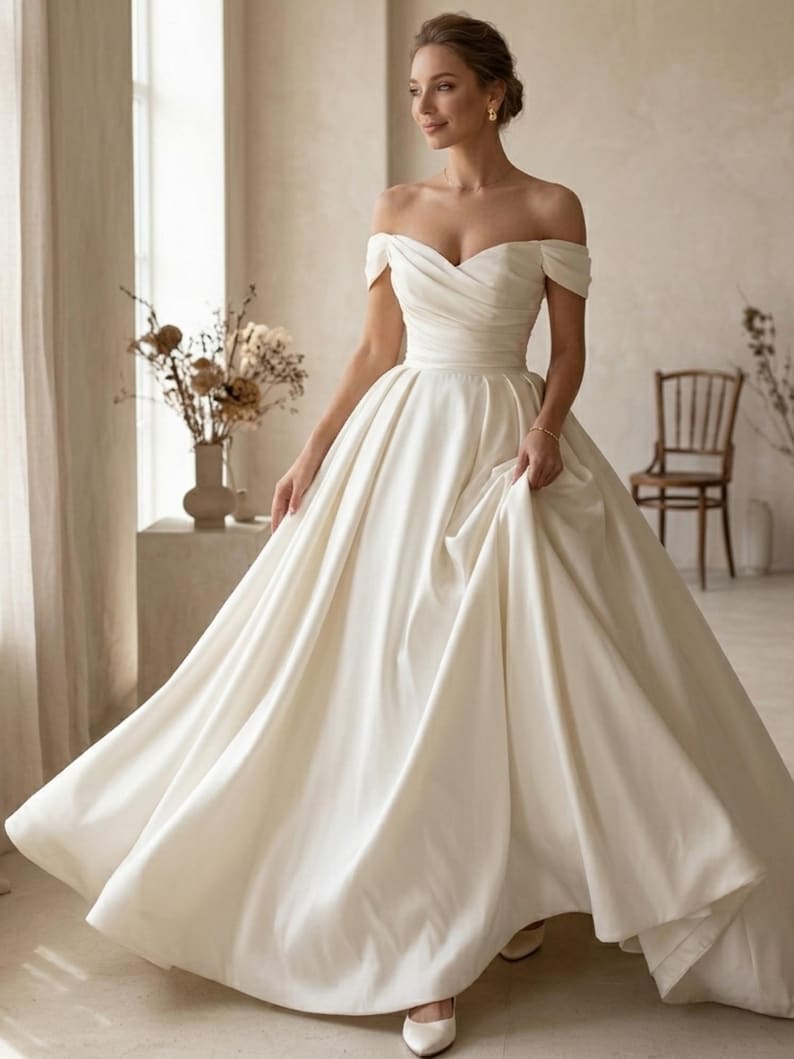 May include: An off-white, off-the-shoulder wedding gown with a full skirt. The dress features a sweetheart neckline and a fitted bodice that flows into a voluminous skirt. The bride is wearing white heels.