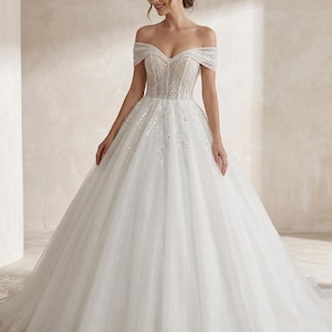 Op de afbeelding: Een witte off-the-shoulder trouwjurk met een sprankelend lijfje en een volle, vloeiende rok. De jurk heeft delicate kralen en een subtiele glans door de stof, wat een elegante en romantische uitstraling creëert. De jurk is ontworpen voor een speciale gelegenheid.