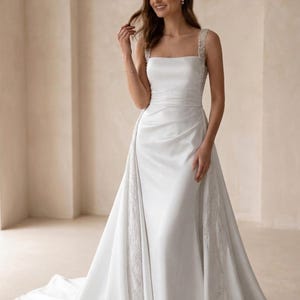 Pode incluir: Um vestido de noiva branco de cetim com decote quadrado e cauda fluida. O vestido apresenta delicados enfeites de contas nas al&ccedil;as e ao longo da lateral da saia. O vestido &eacute; longo e tem um corpete ajustado.
