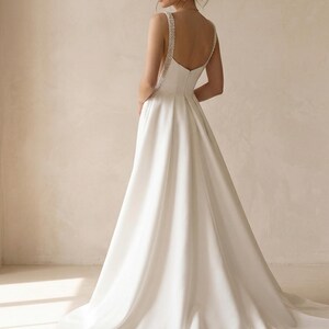 Pode incluir: Um vestido de noiva branco com uma saia fluida e costas baixas. As al&ccedil;as s&atilde;o adornadas com pequenas contas brilhantes. O vestido tem um design simples e elegante, ideal para uma cerim&oacute;nia de casamento.