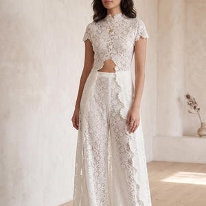 Può includere: Tuta in pizzo bianco con collo alto e maniche corte. La parte superiore ha uno scollo a goccia e i pantaloni a gamba larga hanno un bordo smerlato. L'intricato motivo floreale in pizzo copre l'intero capo.