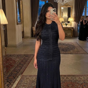 Può includere: Un abito blu navy a due pezzi con un top abbottonato e una gonna abbinata. L'abito ha un design a maglia strutturata. La foto è scattata in una stanza con un grande specchio e un lampadario.