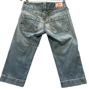 May include: Light blue denim capri pants featuring a mid-rise waist, two back pockets with a signature horseshoe stitch design, and a red brand label on the waistband. The denim has a faded wash, frayed pocket edges, and wide hemmed cuffs.