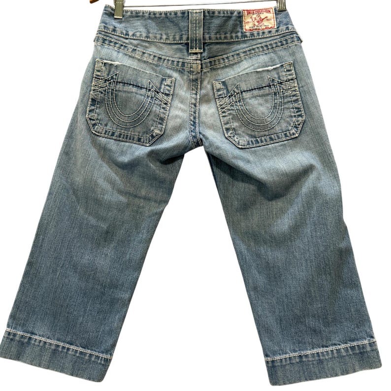 May include: Light blue denim capri pants featuring a mid-rise waist, two back pockets with a signature horseshoe stitch design, and a red brand label on the waistband. The denim has a faded wash, frayed pocket edges, and wide hemmed cuffs.