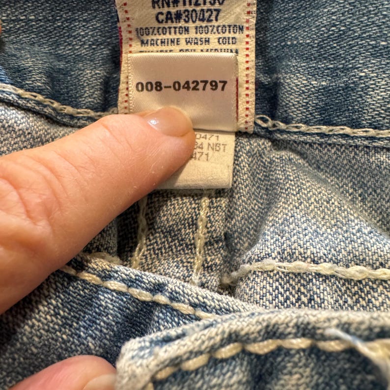 May include: Light blue denim fabric featuring thick white contrast stitching. A white interior garment tag displays the text RN#112790, CA#30427, 100% COTTON 100% COTON, MACHINE WASH COLD, and the numerical sequence 008-042797, along with additional smaller text.
