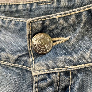 May include: Light blue denim jeans featuring thick, cream-colored contrast stitching and a silver-toned metal button embossed with the text True Religion Brand Jeans and a horseshoe logo. The fabric has a faded, washed-out appearance.