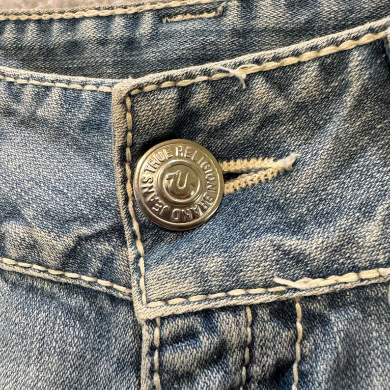 May include: Light blue denim jeans featuring thick, cream-colored contrast stitching and a silver-toned metal button embossed with the text True Religion Brand Jeans and a horseshoe logo. The fabric has a faded, washed-out appearance.