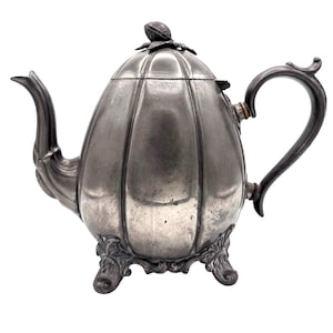 May include: A metallic gray teapot with a ribbed, pumpkin-like body, a curved spout, and a handle. The lid features a small decorative finial, and the base is supported by three ornate, scrolled feet. The surface has a dull, aged finish.