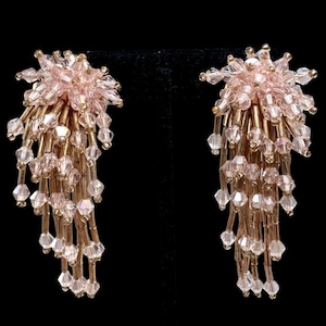 May include: A pair of chandelier earrings featuring cascading strands of light pink faceted crystal beads and elongated metallic bugle beads. The design includes a cluster of pink crystals at the top, with multiple dangling beaded fringe elements.