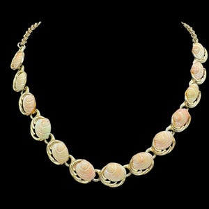May include: A gold-tone metal necklace featuring a series of oval-shaped, light pink and cream-coloured seashell charms. Each shell is encased in a decorative gold-tone metal frame, connected by small circular links to form a continuous chain.