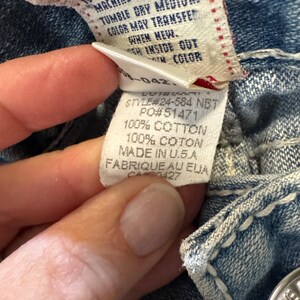 May include: A blue denim garment featuring a white interior care label with black text. The label displays Style #24-584 NBT, PO #51471, and states 100% Cotton, Made in U.S.A. The fabric shows thick white contrast stitching along the seams.
