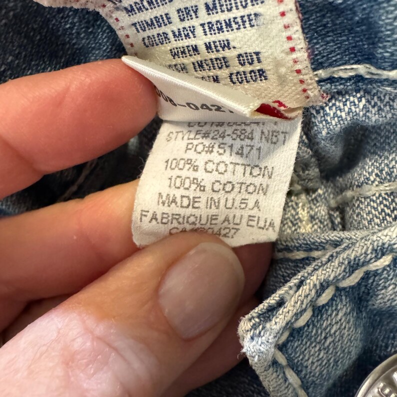 May include: A blue denim garment featuring a white interior care label with black text. The label displays Style #24-584 NBT, PO #51471, and states 100% Cotton, Made in U.S.A. The fabric shows thick white contrast stitching along the seams.